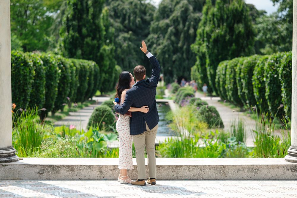 Couple embracing in a formal garden with trimmed hedges, columns, and a reflecting pool on a sunny day.
