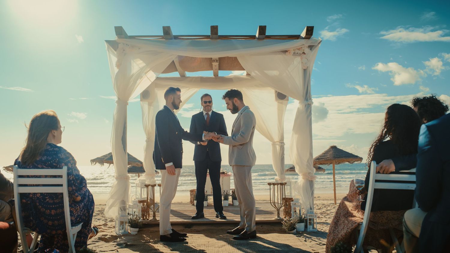 Two gold wedding rings on a sandy beach with a couple kissing by the ocean in the background.