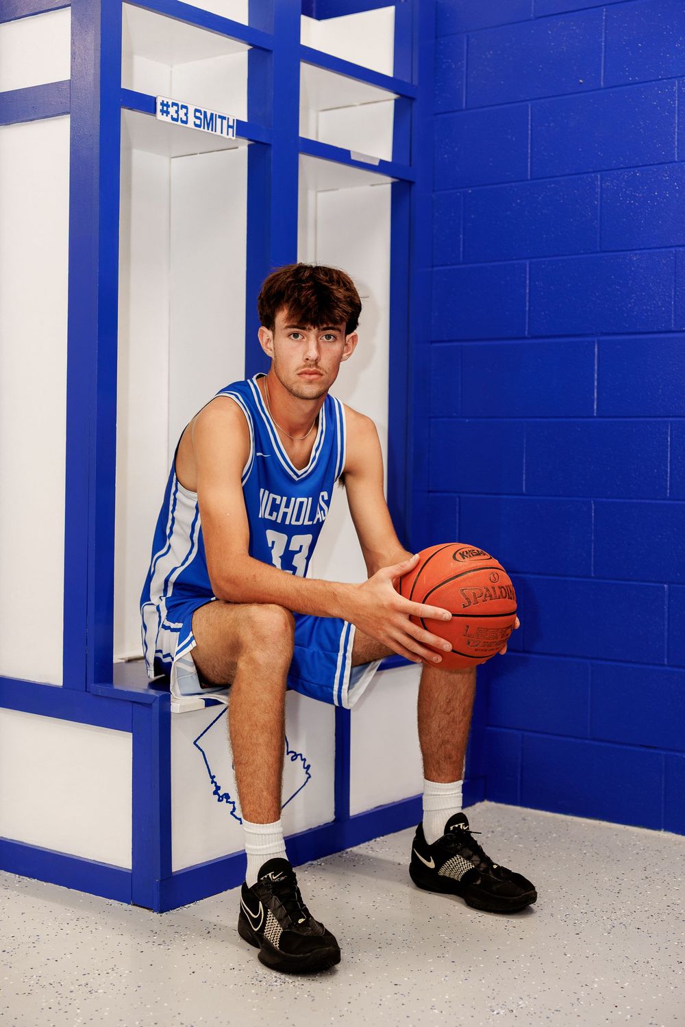 Basketball player #33 Smith sits in a locker room holding a Spalding ball, wearing a blue Nichols uniform and Nike shoes.