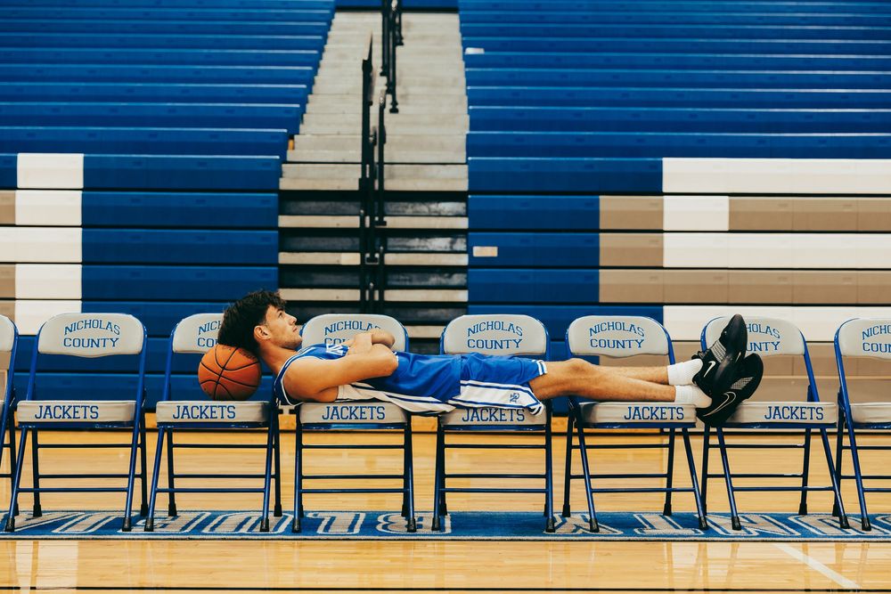 Basketball player resting on gym chairs at Murray County Jackets indoor court with blue bleachers.