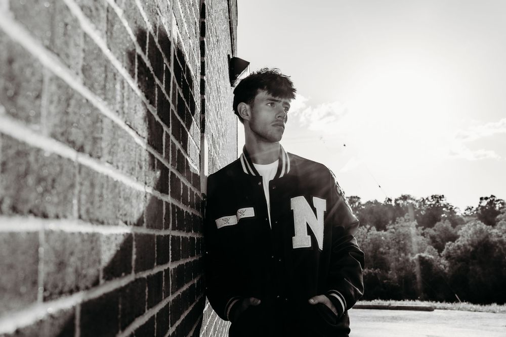 Young man in varsity jacket leans against brick wall in black and white portrait photo outdoors.