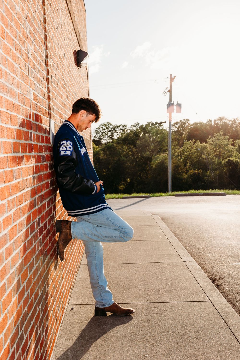 Young man in varsity jacket and jeans leaning against brick wall outdoors, looking down in golden sunlight.