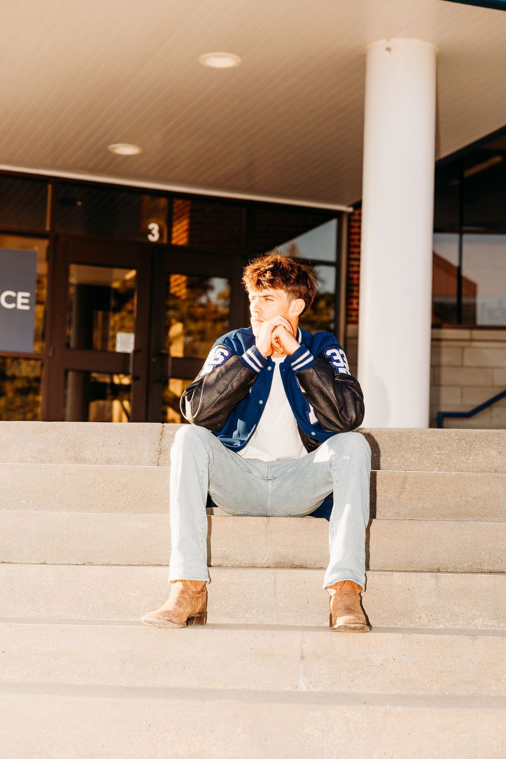 Young man in blue varsity jacket and light jeans sitting on concrete steps outside a building.