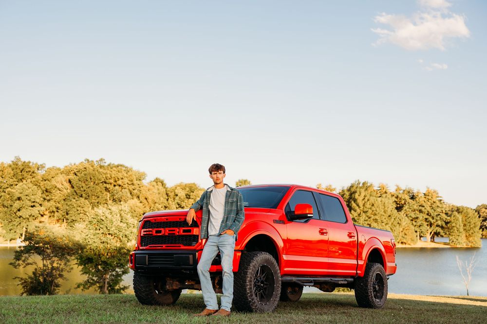 Young man leaning against a red Ford F-150 Raptor truck parked by a lake with trees in background.