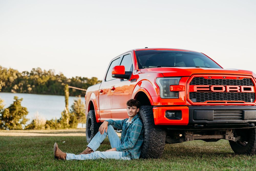 A young woman in a denim jacket sits on grass beside a red Ford F-150 Raptor truck near a lake.