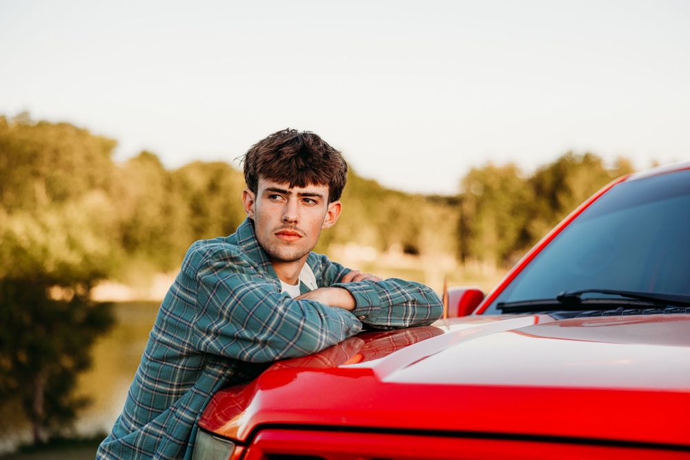 Young man in plaid shirt leaning on red car hood outdoors near trees at golden hour.