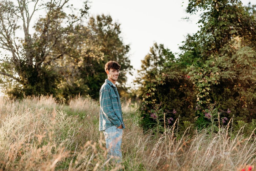 Person standing in a sunlit field with tall grass and green trees in the background during golden hour.