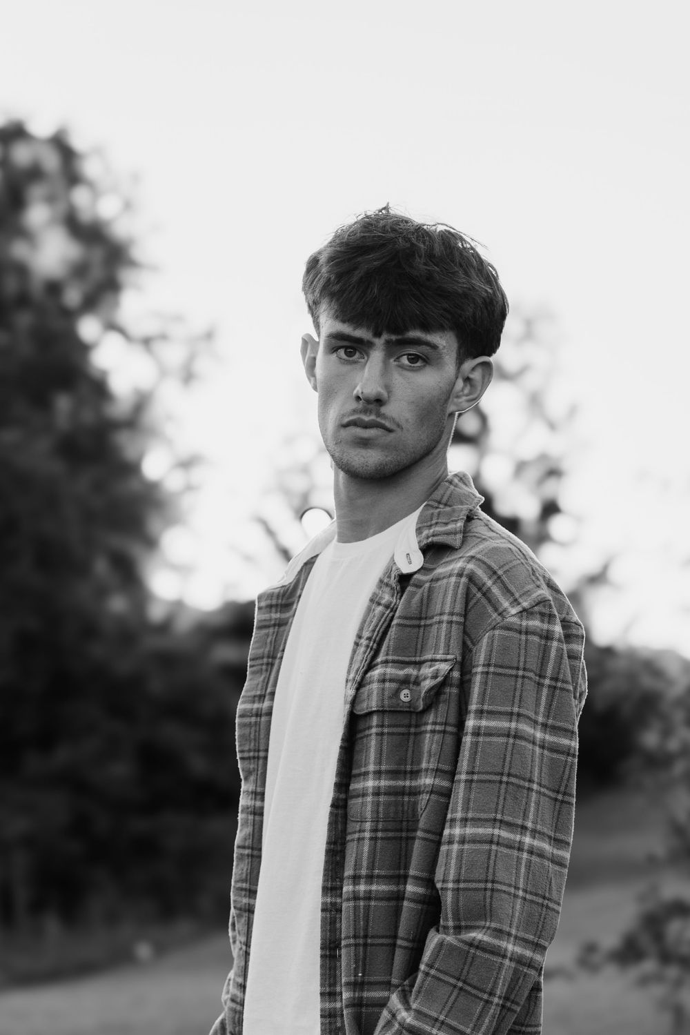 Black and white portrait of a young man in a plaid shirt and white tee, standing outdoors with trees in background.