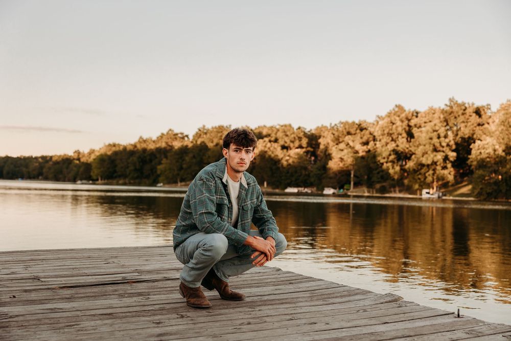 Young man crouching on a wooden dock by a calm lake at golden hour with autumn trees in the background.