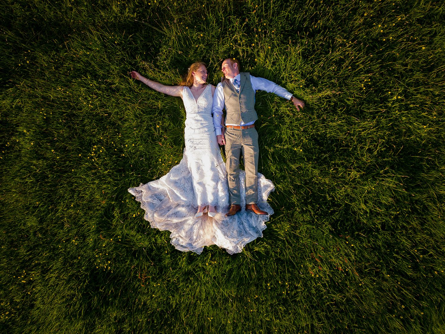 Aerial drone view of bride and groom in wedding attire lying on lush green grass, arms outstretched.