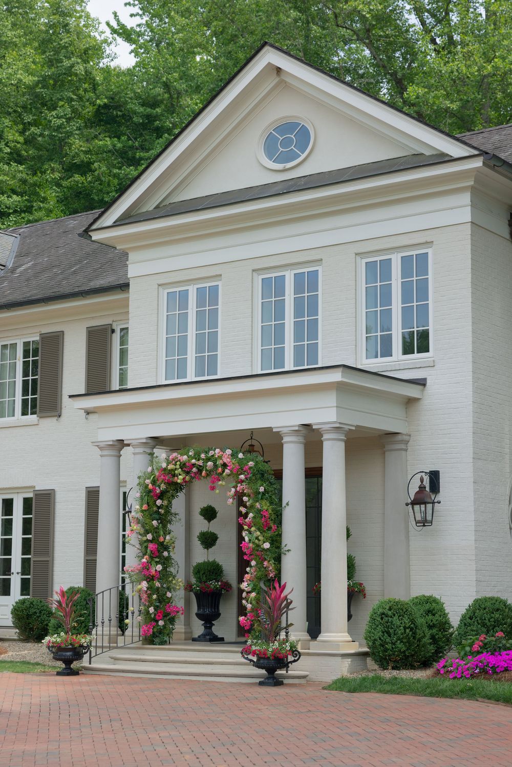 White colonial mansion with floral arch entrance, columns, and manicured shrubs on brick driveway.