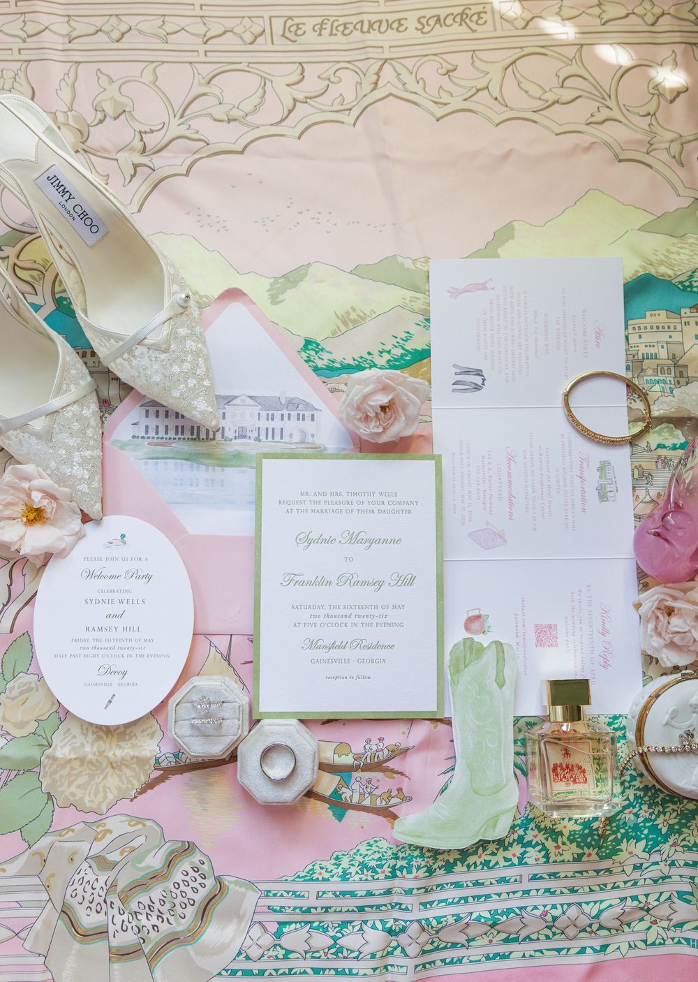 Elegant wedding invitation suite with floral details, gold rings, perfume, and Jimmy Choo heels on a pink illustrated map.