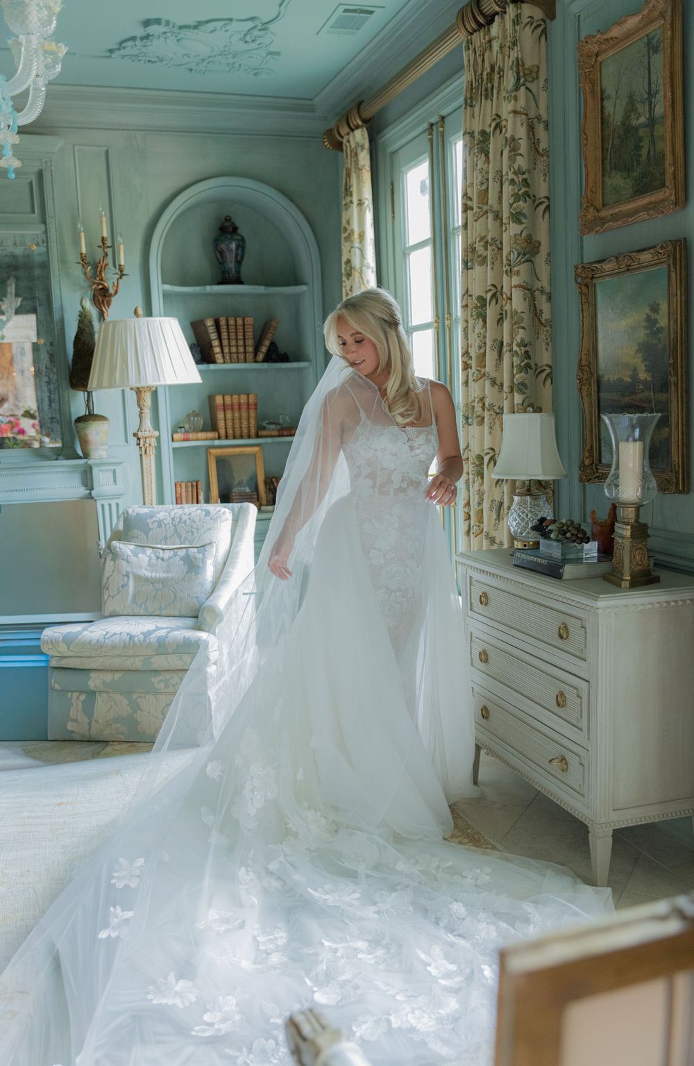 Bride in elegant white lace wedding gown with long veil in a luxurious blue French-style room.