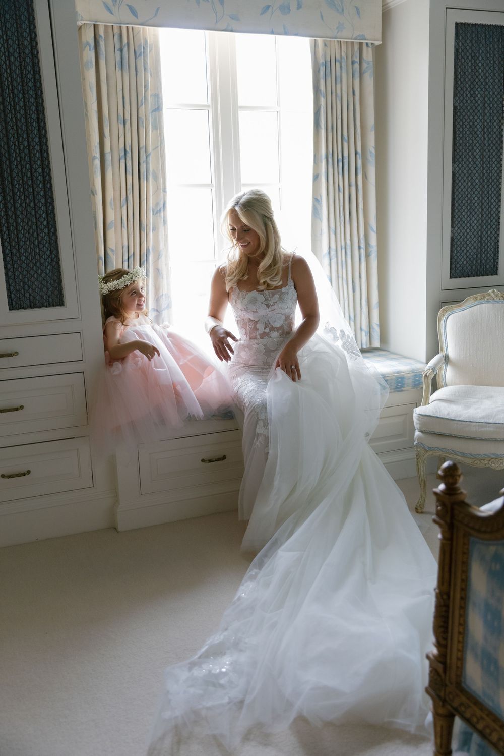 Bride in white lace gown with flower girl in pink tulle dress by sunlit window in elegant room.