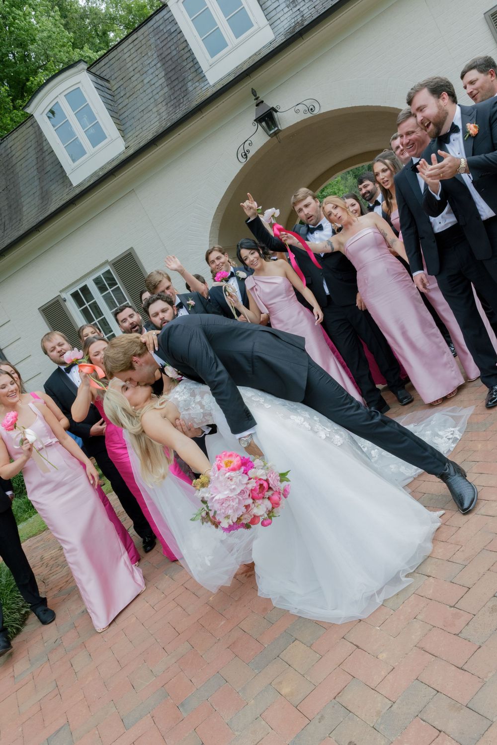 Groom dips bride in white gown surrounded by cheering wedding party with pink bridesmaid dresses outdoors.