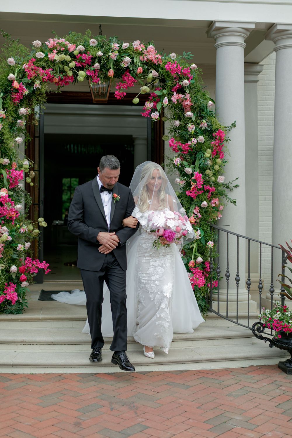 Bride and groom exit a venue adorned with a lush pink and green floral arch on a brick staircase.