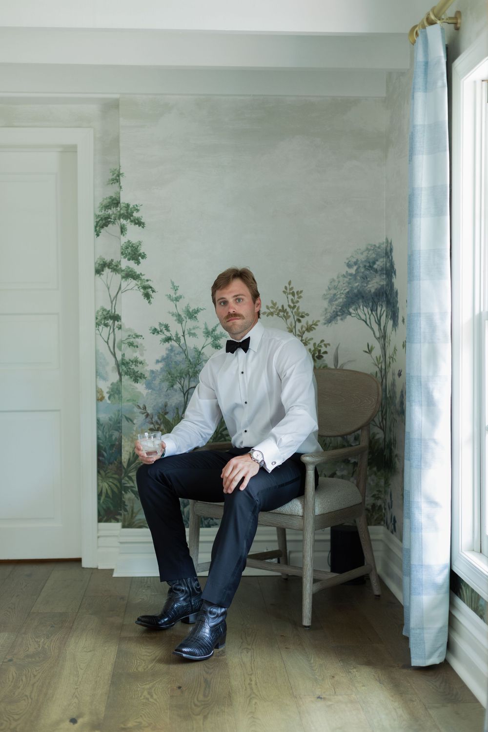 A well-dressed man in a tuxedo shirt and bow tie sits in a chair holding a drink, posed before an elegant scenic wallpaper.