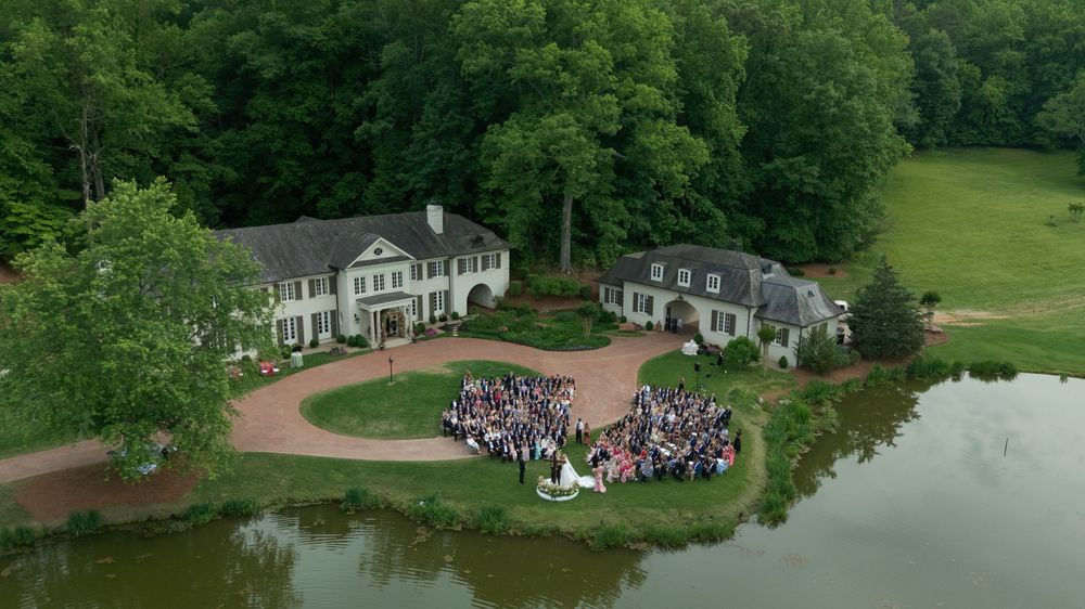 Aerial view of a large wedding gathering on lush green estate grounds beside a pond, featuring an elegant white mansion.