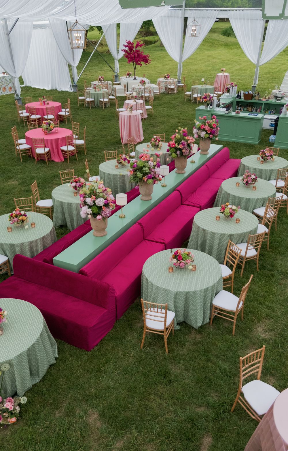 Elegant outdoor wedding reception with fuchsia sofas, mint green tables, gold chairs, and pink floral centerpieces under a white tent.