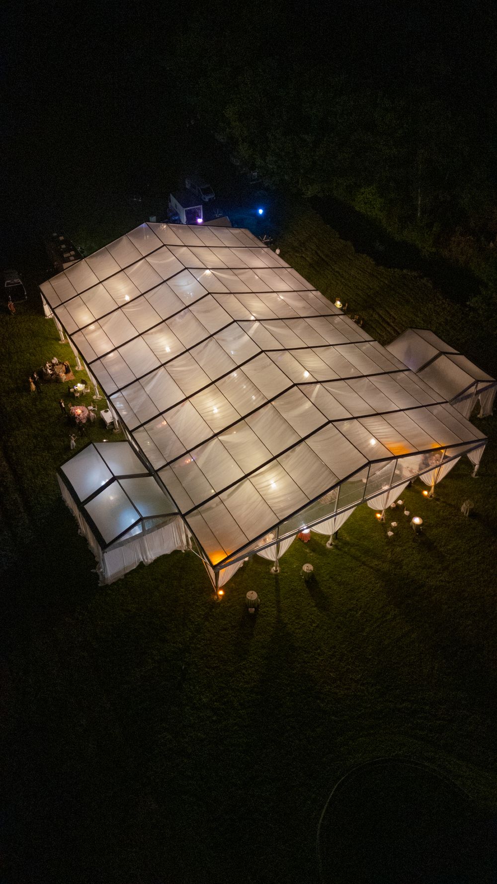 Aerial night view of illuminated clear marquee tent on green lawn with warm glowing lights at outdoor event.
