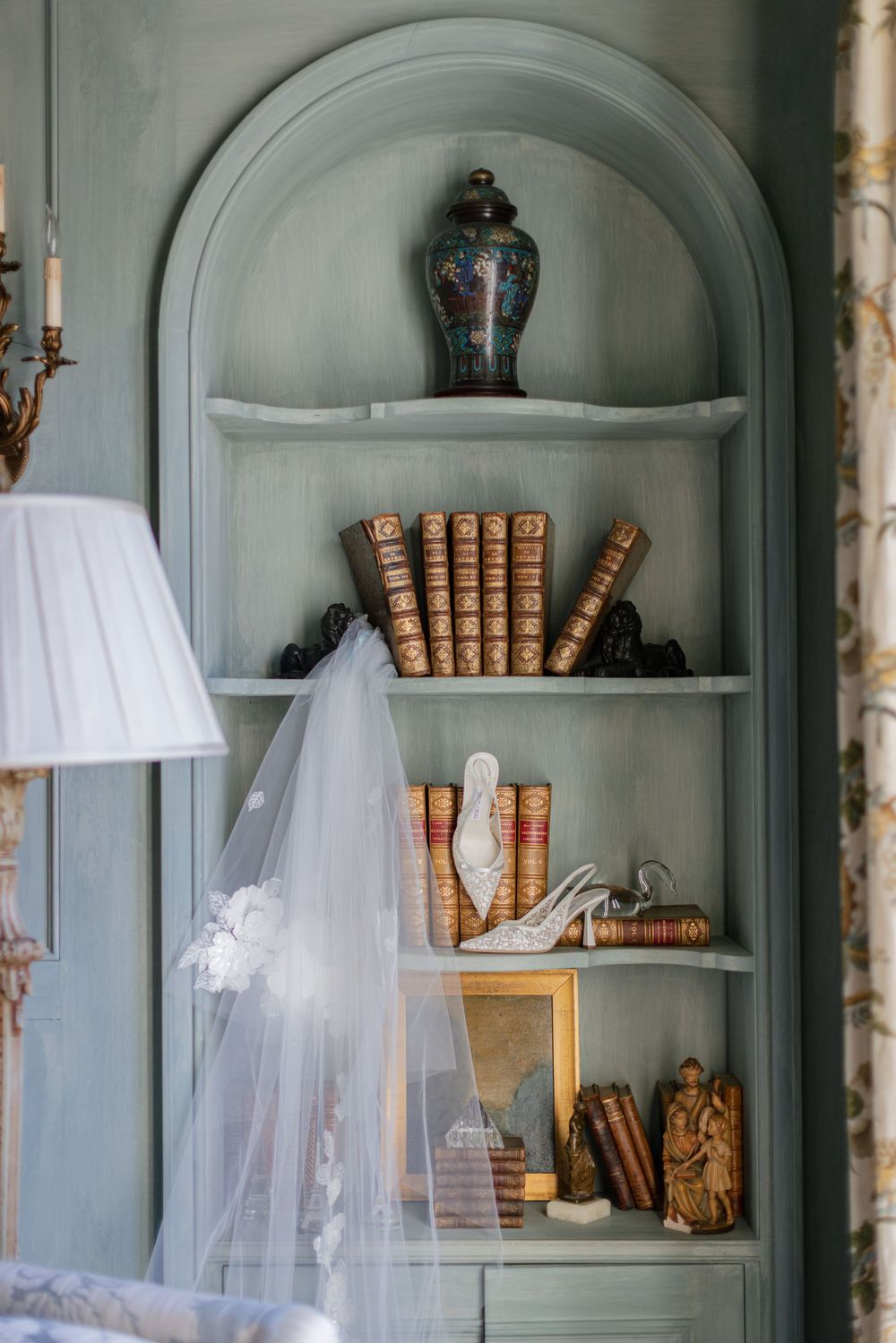 Bridal veil draped over a vintage bookshelf with antique books, decorative vase, and wedding heels in a blue-gray alcove.