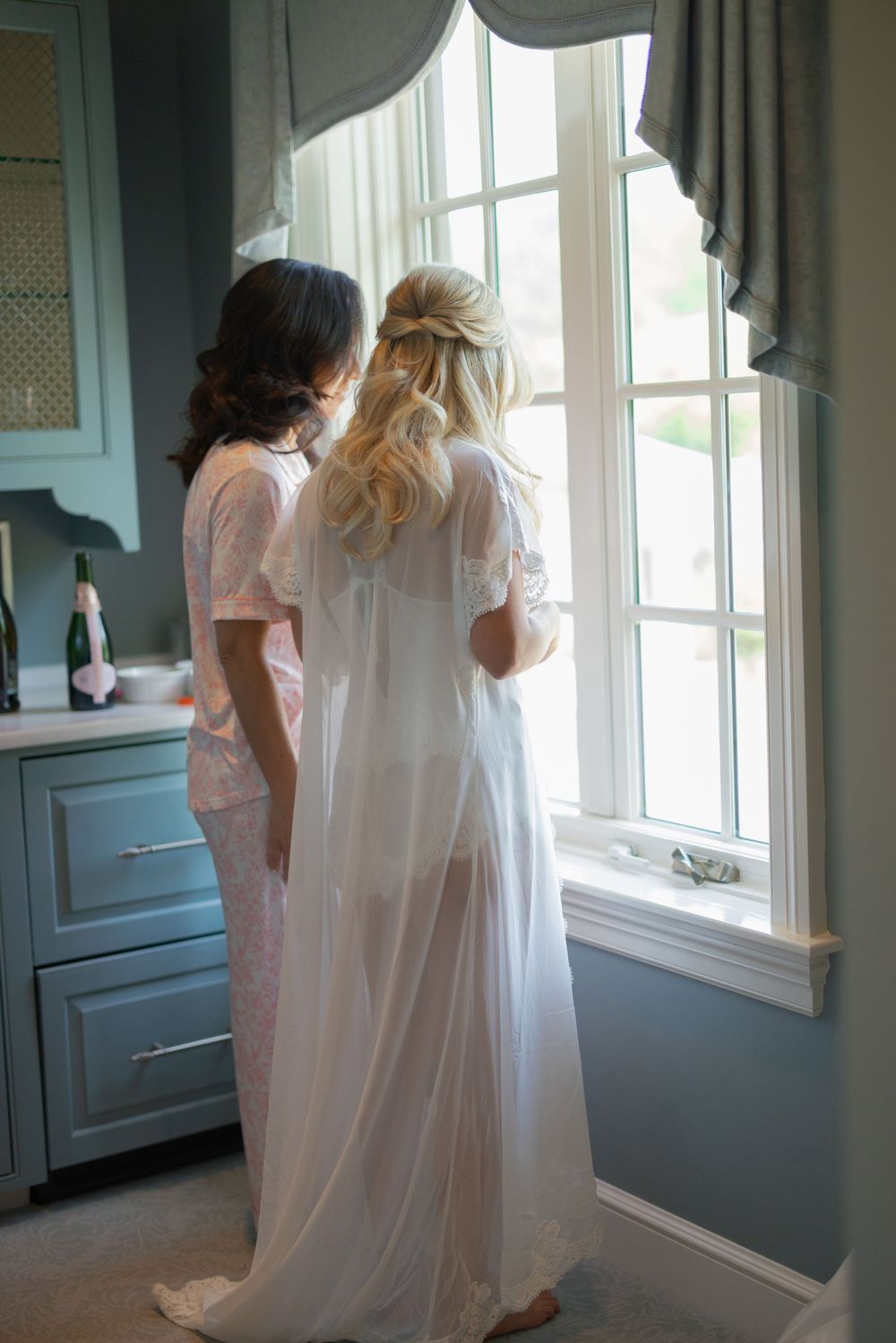 Two women in robes stand by a bright window, one in white sheer bridal robe, sharing a quiet moment.