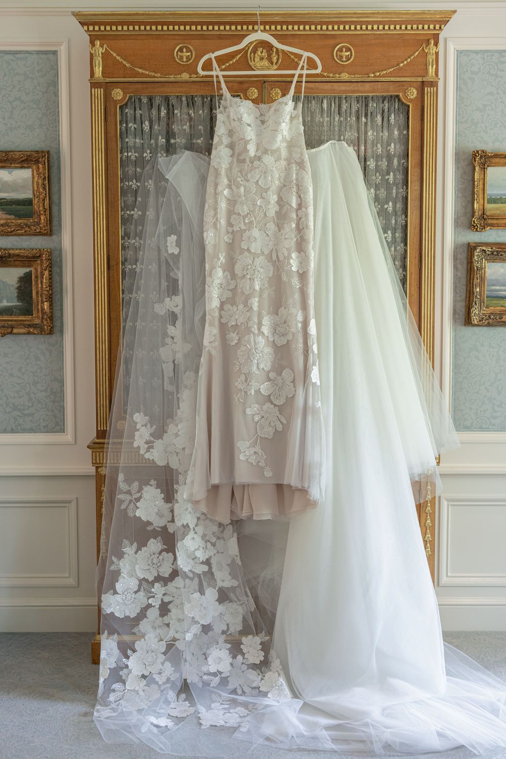 Elegant lace wedding dress with floral appliques and tulle veil displayed on a gold ornate mirror frame.