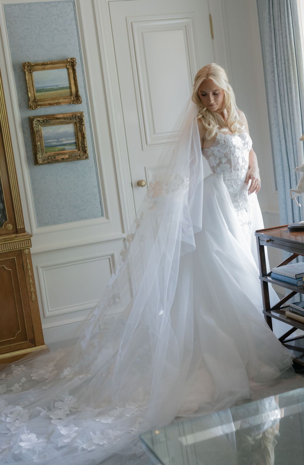 Blonde bride in elegant white ball gown with lace details and flowing veil in a luxurious room.