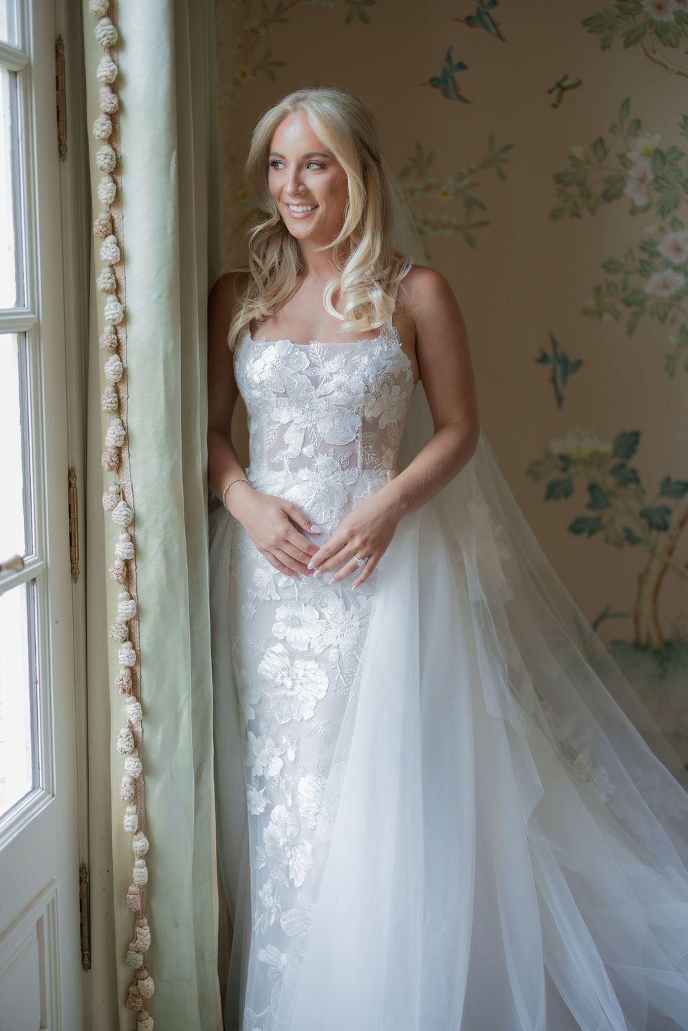 Smiling bride in floral lace wedding gown with tulle overlay stands by window in elegant floral wallpapered room.