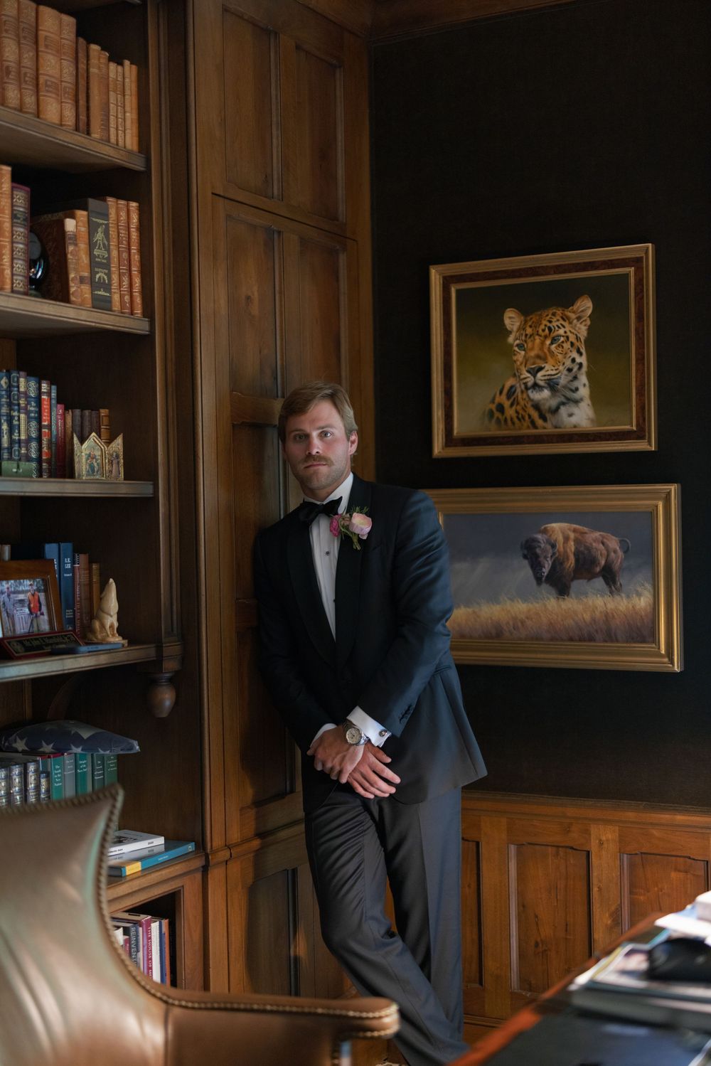 Groom in tuxedo poses in wood-paneled library with leopard and bison paintings.
