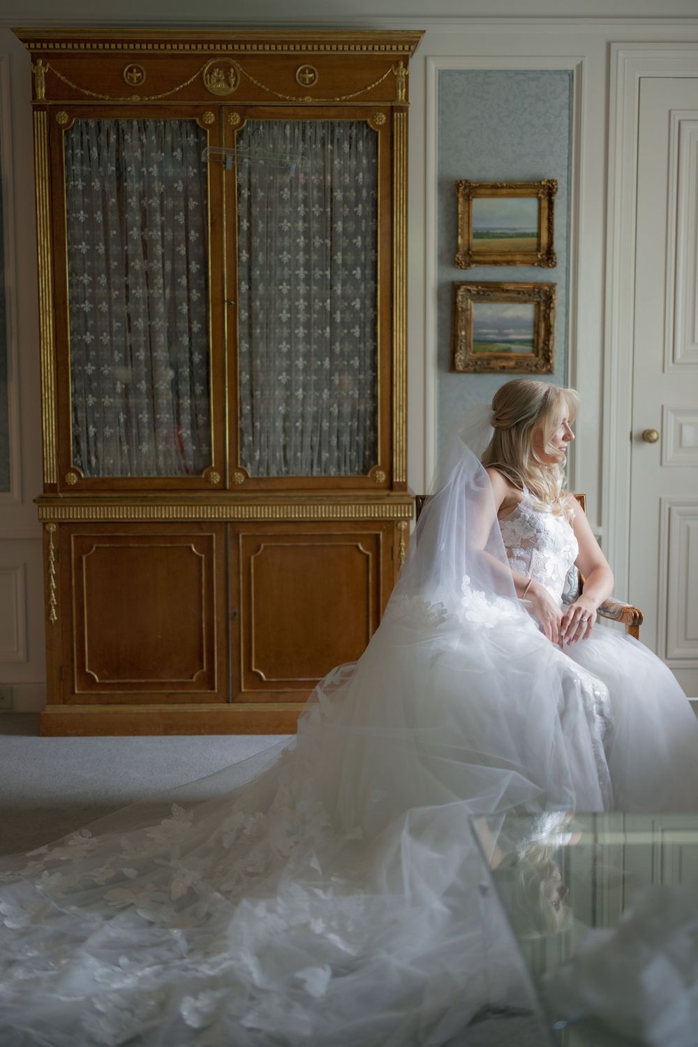 Bride in a flowing white ball gown and veil sits pensively beside an ornate gold cabinet in an elegant room.
