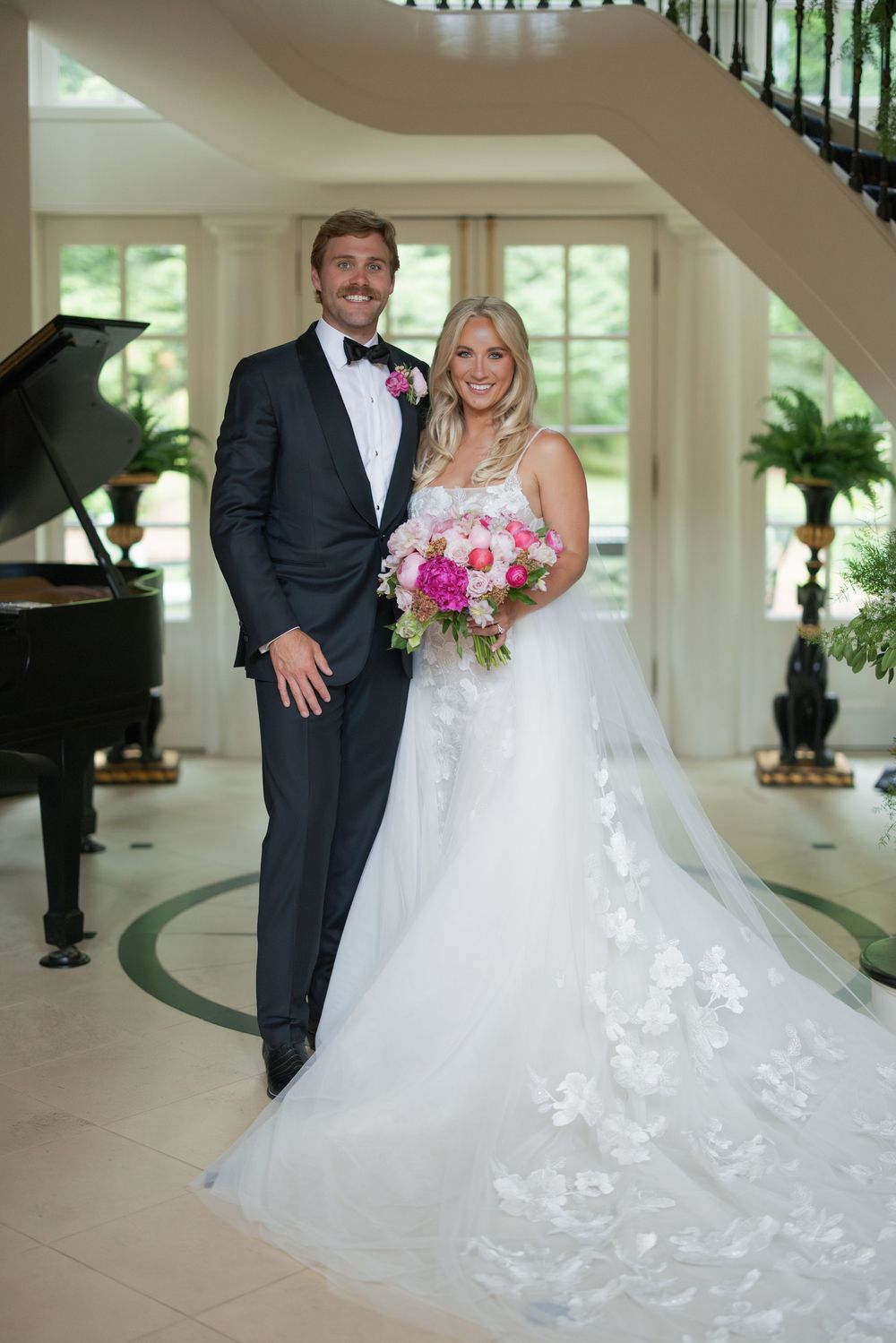 Bride and groom pose elegantly in grand foyer with piano, bride holding vibrant pink bouquet in floral wedding gown.