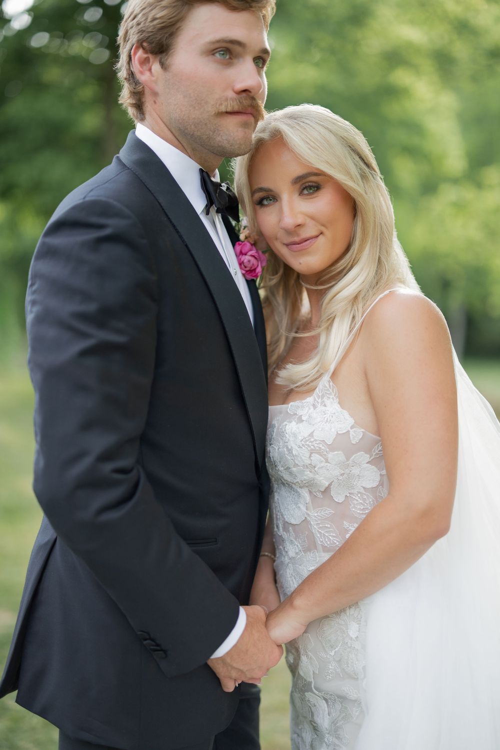 Bride and groom pose together outdoors, she in a floral lace gown, he in a dark suit with pink boutonniere.