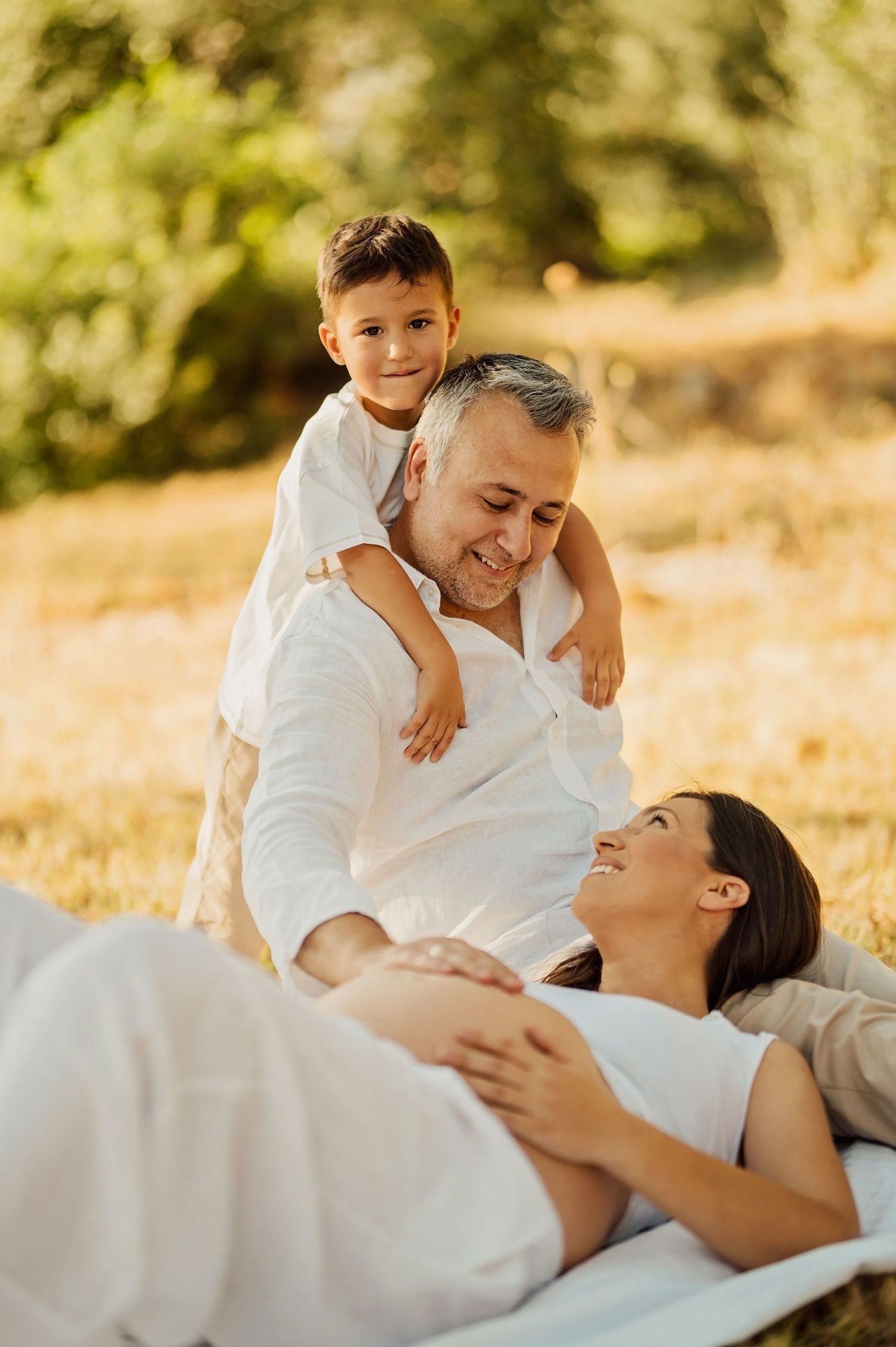 Pregnant woman lying on blanket outdoors with husband and young son in sunny park during family maternity photo session.