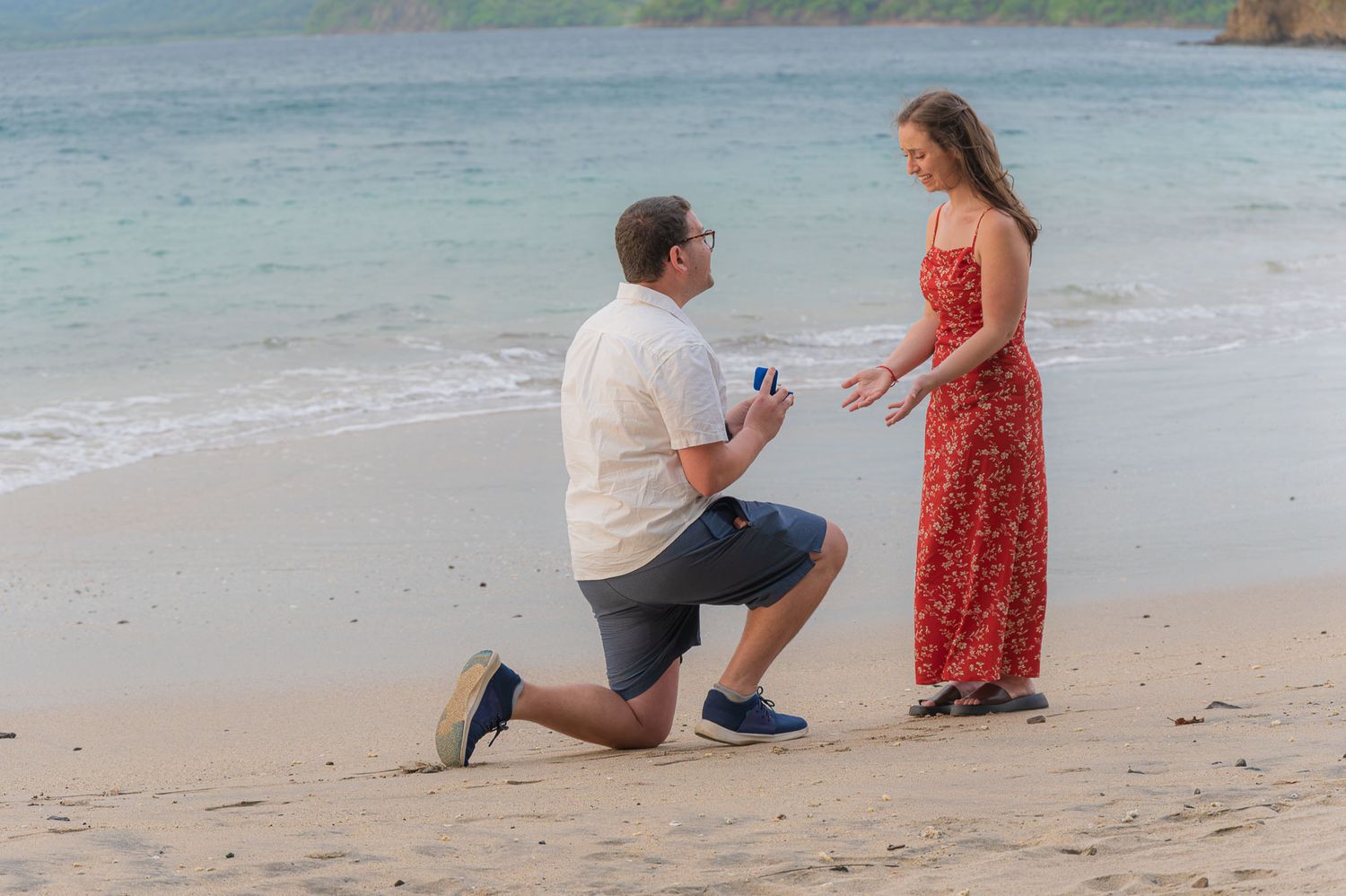 Man proposing on one knee to woman in red dress on tropical beach with ocean waves in background.