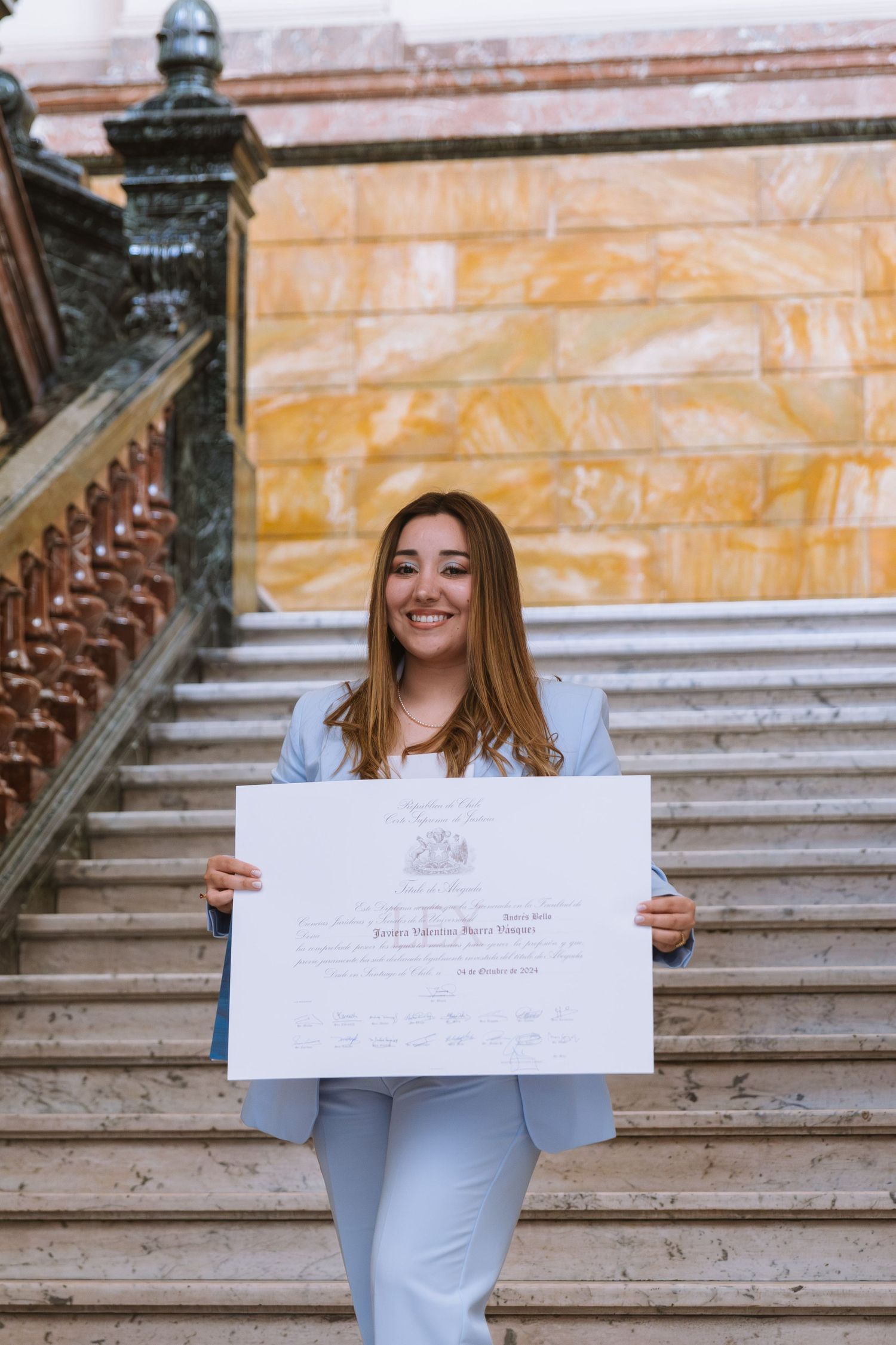 Happy young woman in blue suit proudly displaying her diploma on elegant marble stairs.