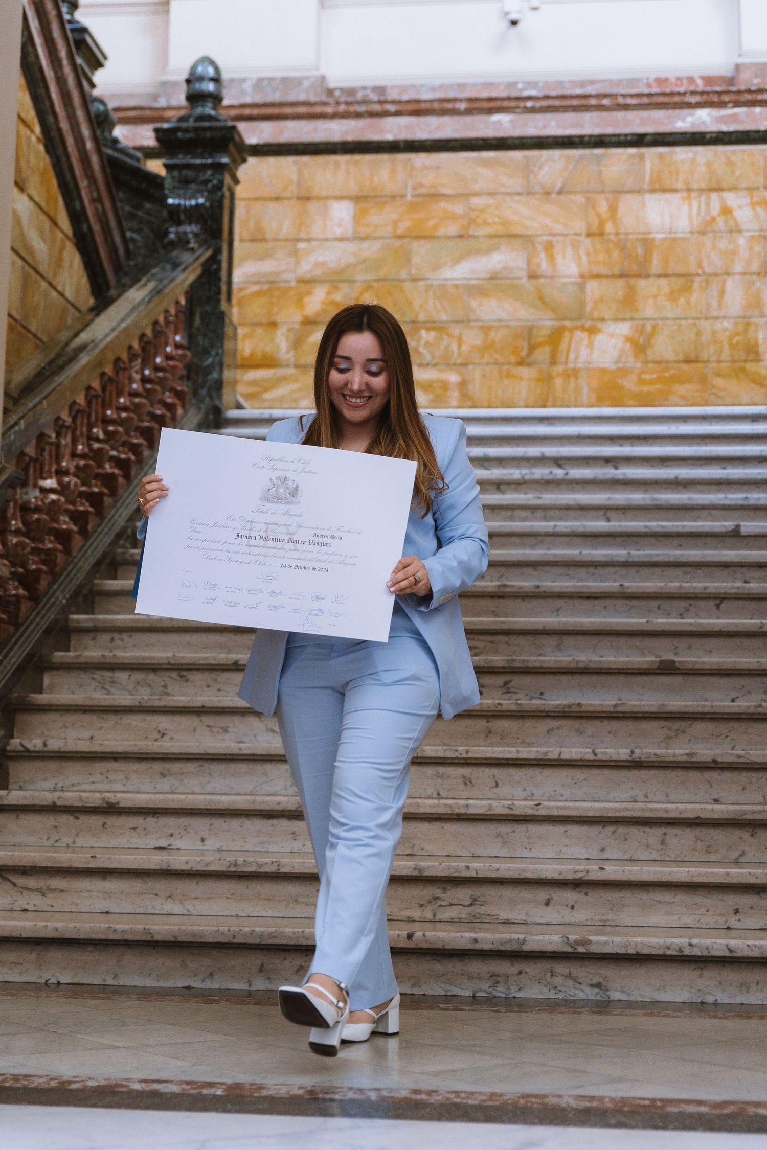 Young woman in light blue suit holding diploma certificate while descending grand marble staircase.