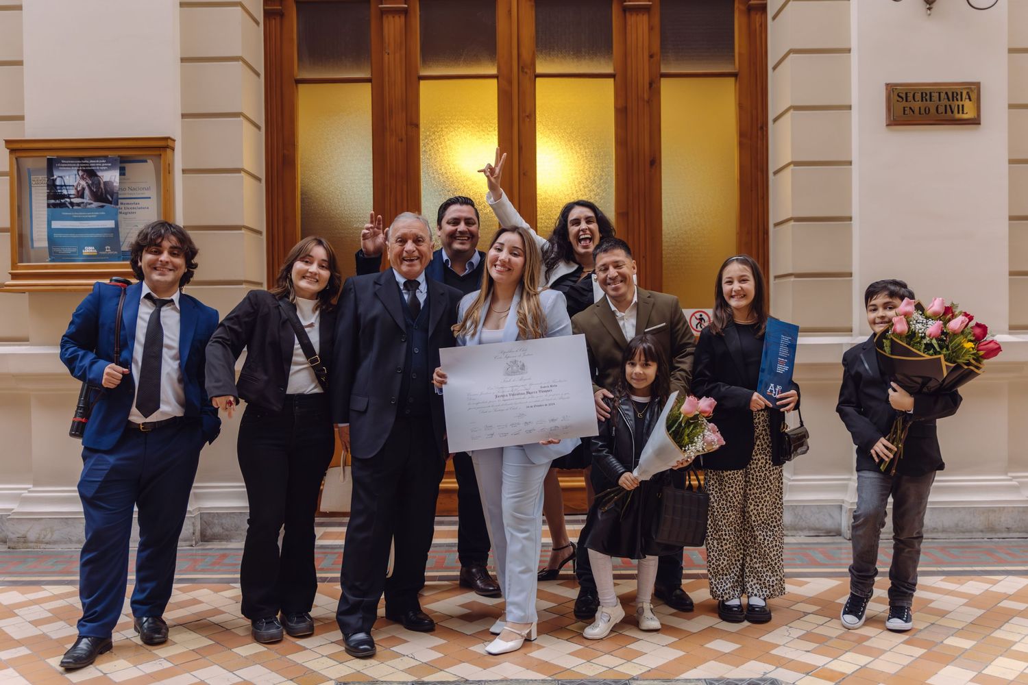 Group of people celebrating graduation with flowers and certificate in elegant building interior.