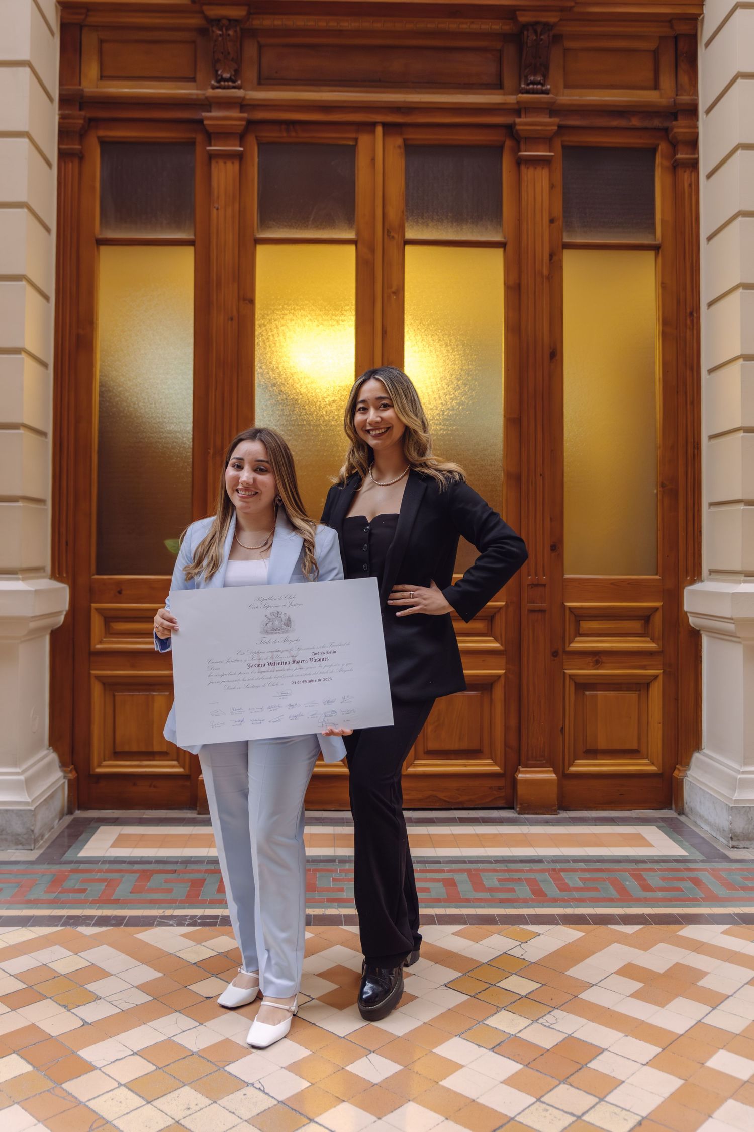 Two young women posing with diploma certificate in front of ornate wooden doors on tiled floor.