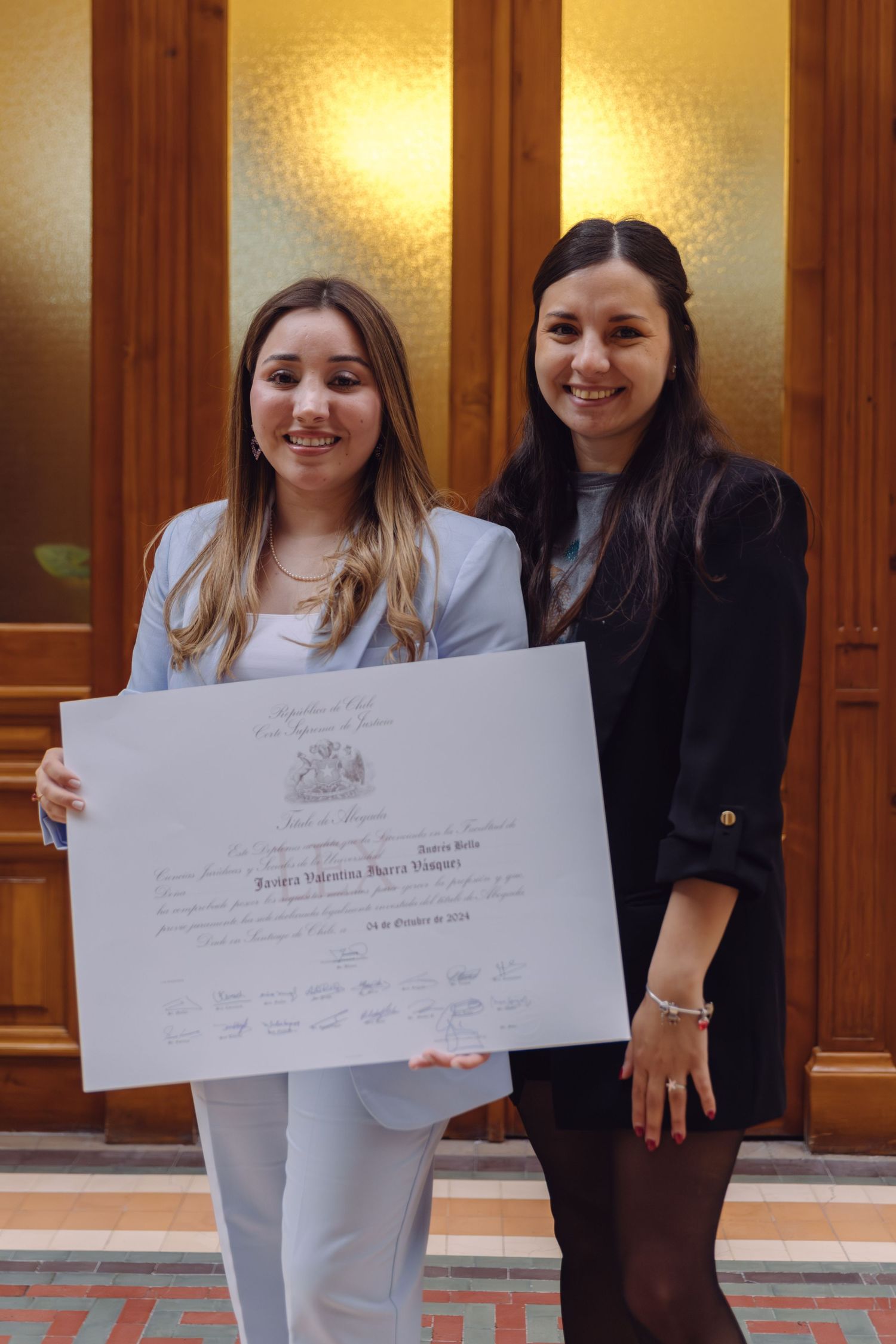 Two smiling young women holding large diploma certificate together in front of wooden doors.