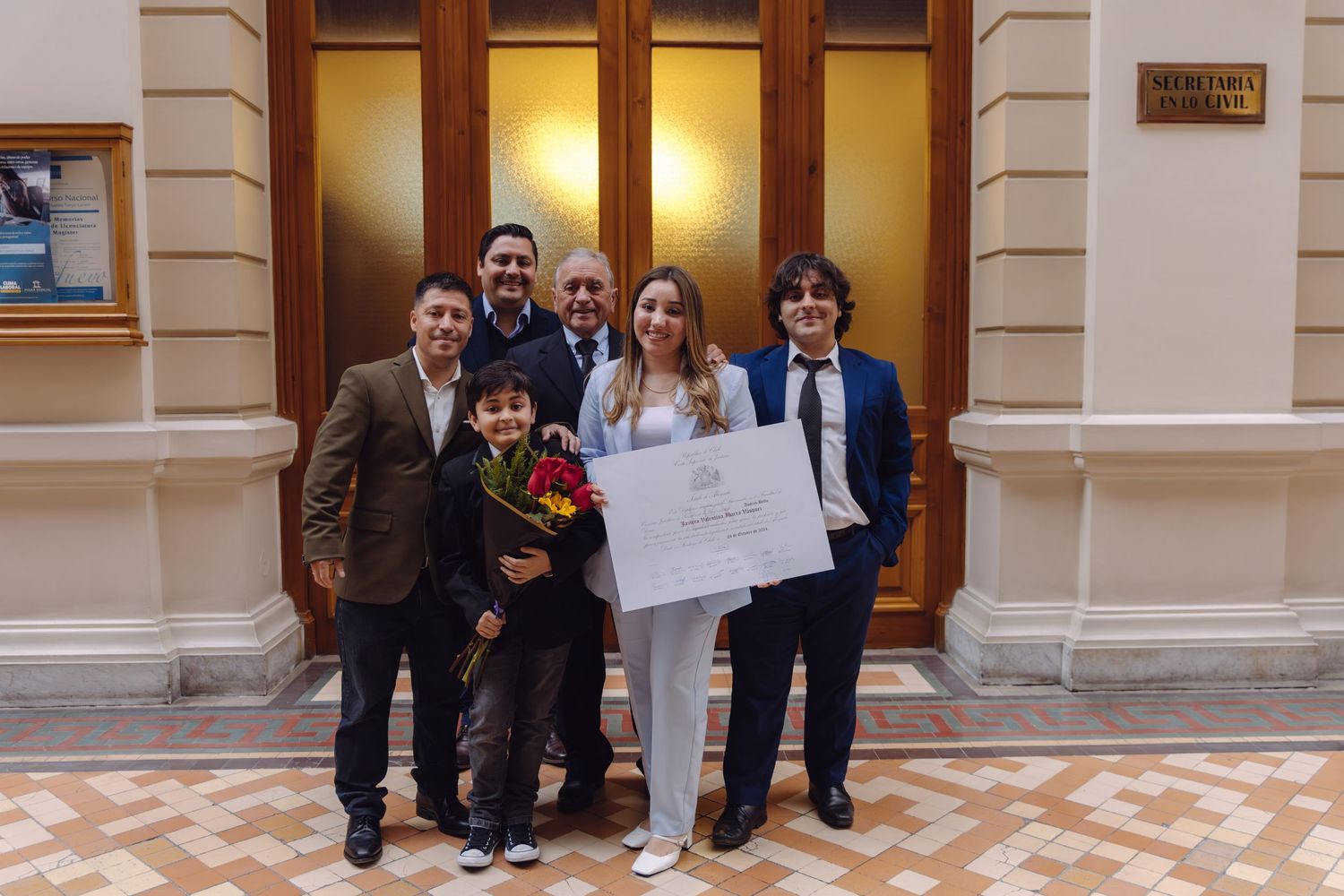 Family group posing with graduation certificate and red roses in elegant building hallway.