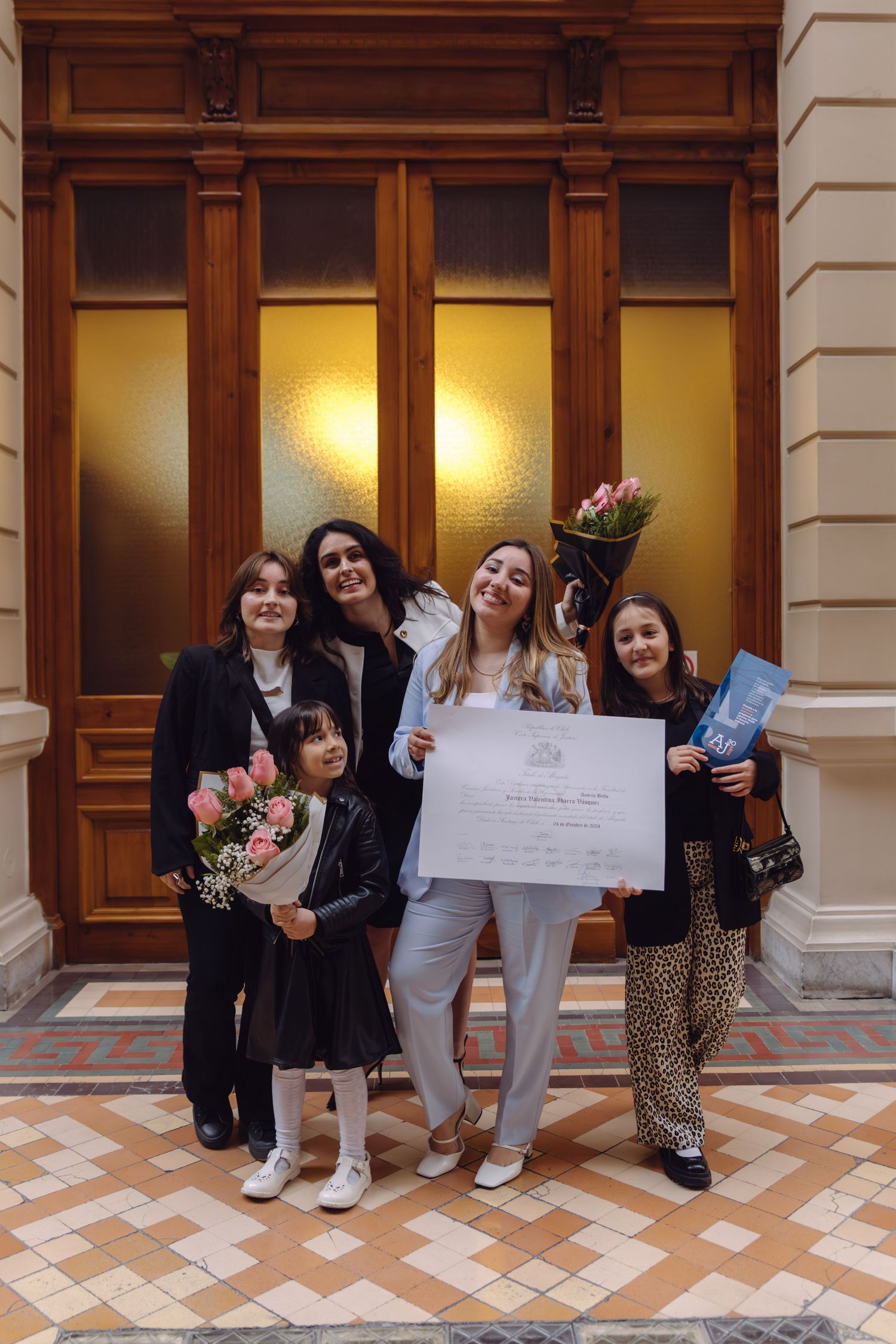 Group of four females posing with diploma certificate and pink flowers in front of ornate doors.