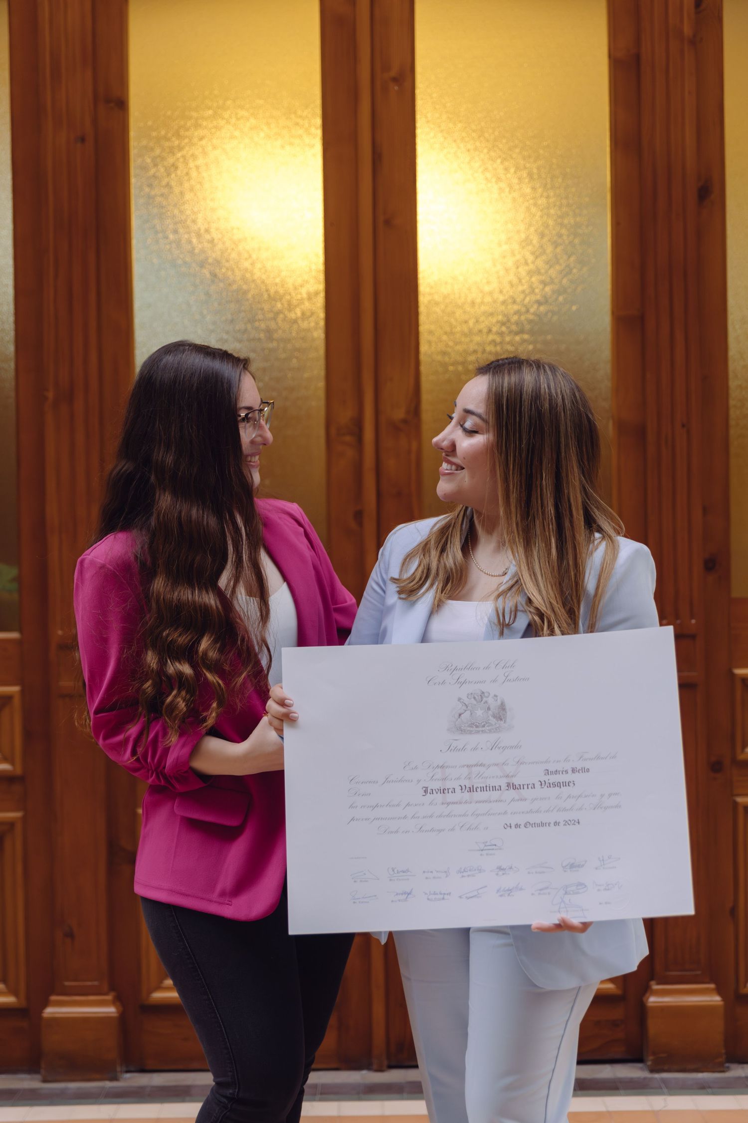 Two women holding diploma certificate together, smiling at each other in front of wooden doors.