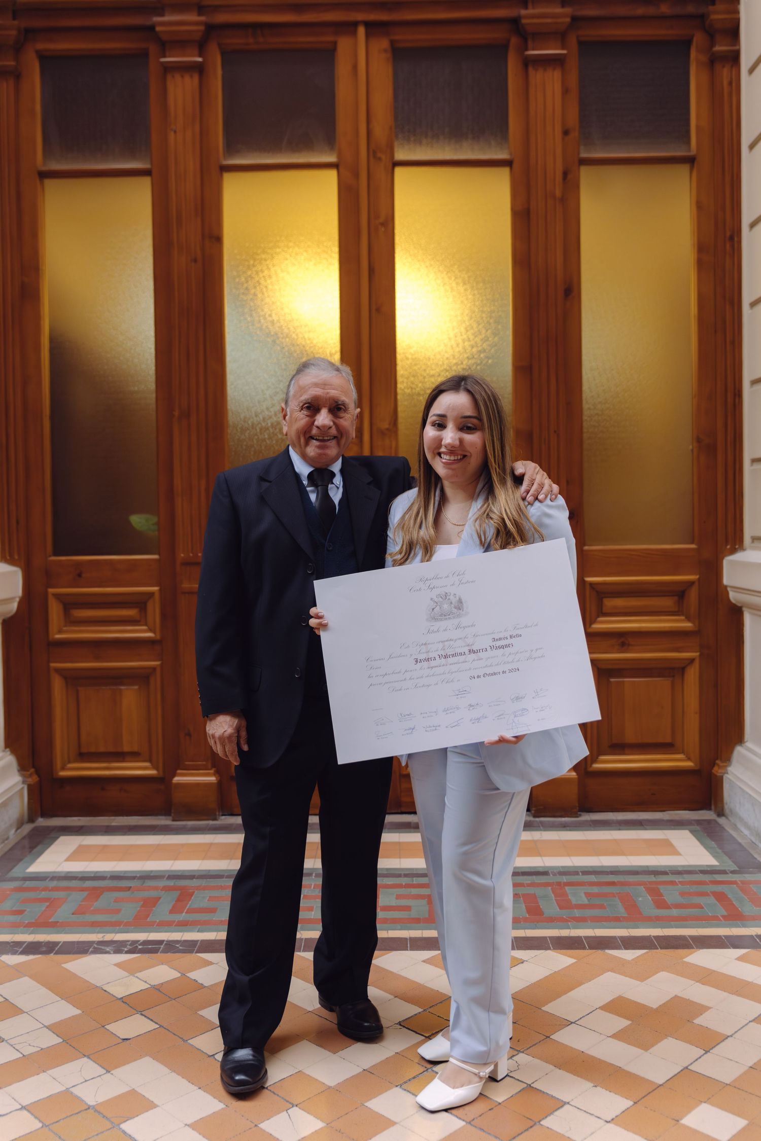 Elderly man and young woman in blue suit holding diploma certificate in front of wooden doors.