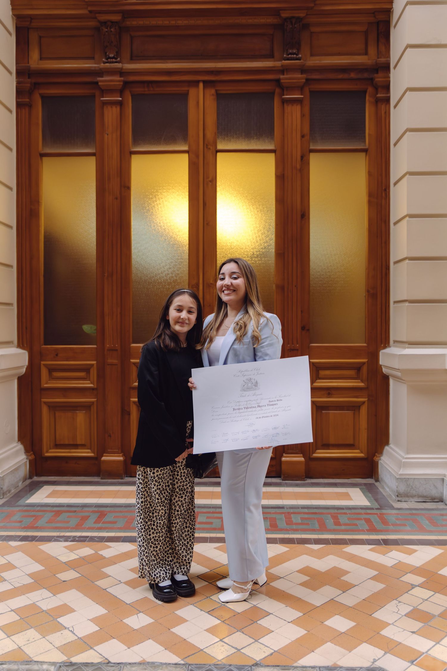 Young girl and woman in blue suit holding diploma certificate in front of ornate wooden doors.