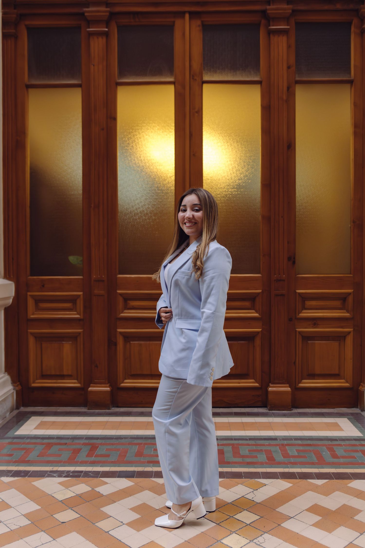 Smiling young woman in light blue pantsuit posing in front of illuminated ornate wooden doors.