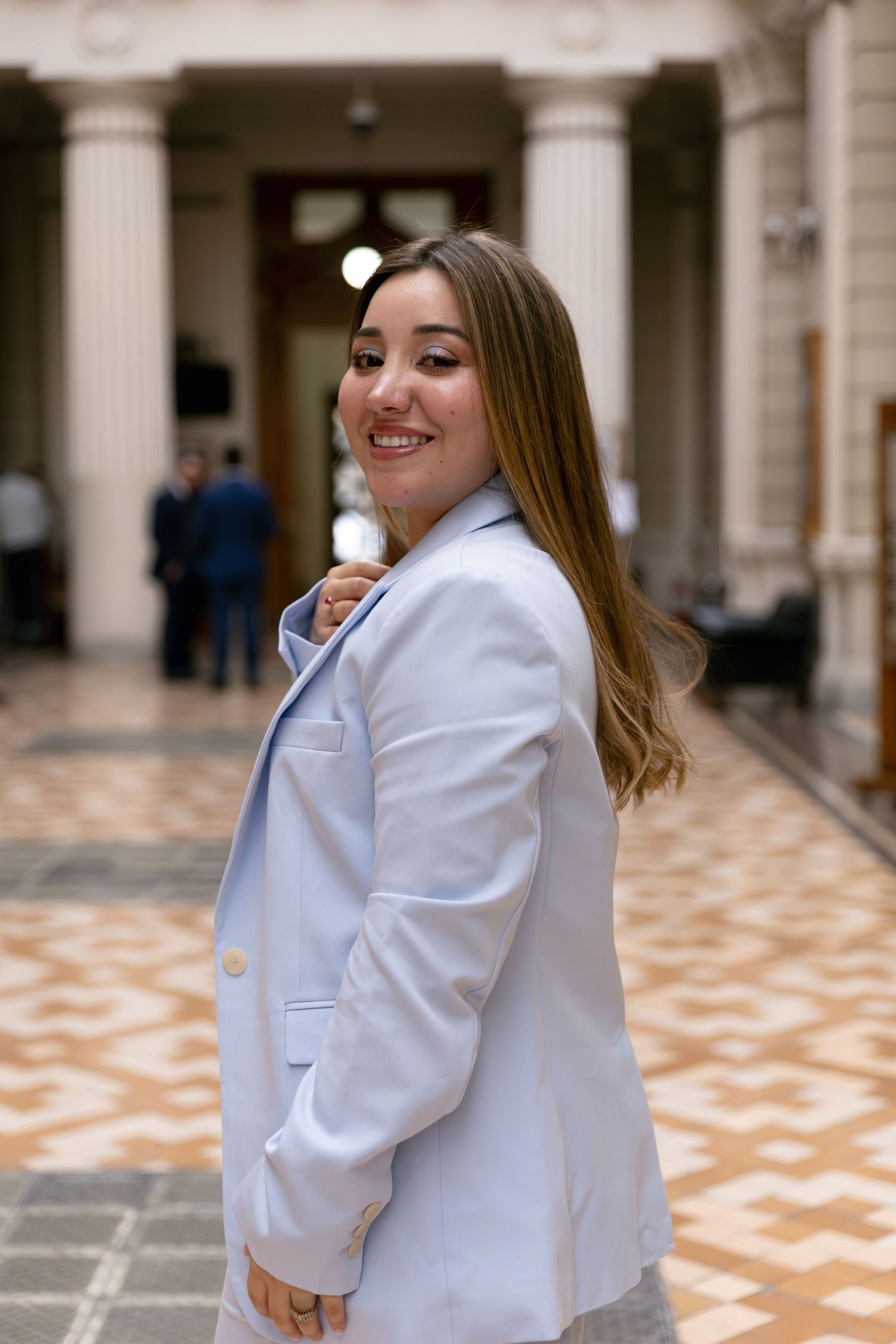 Young woman in light blue blazer smiling over shoulder in elegant columned building interior.