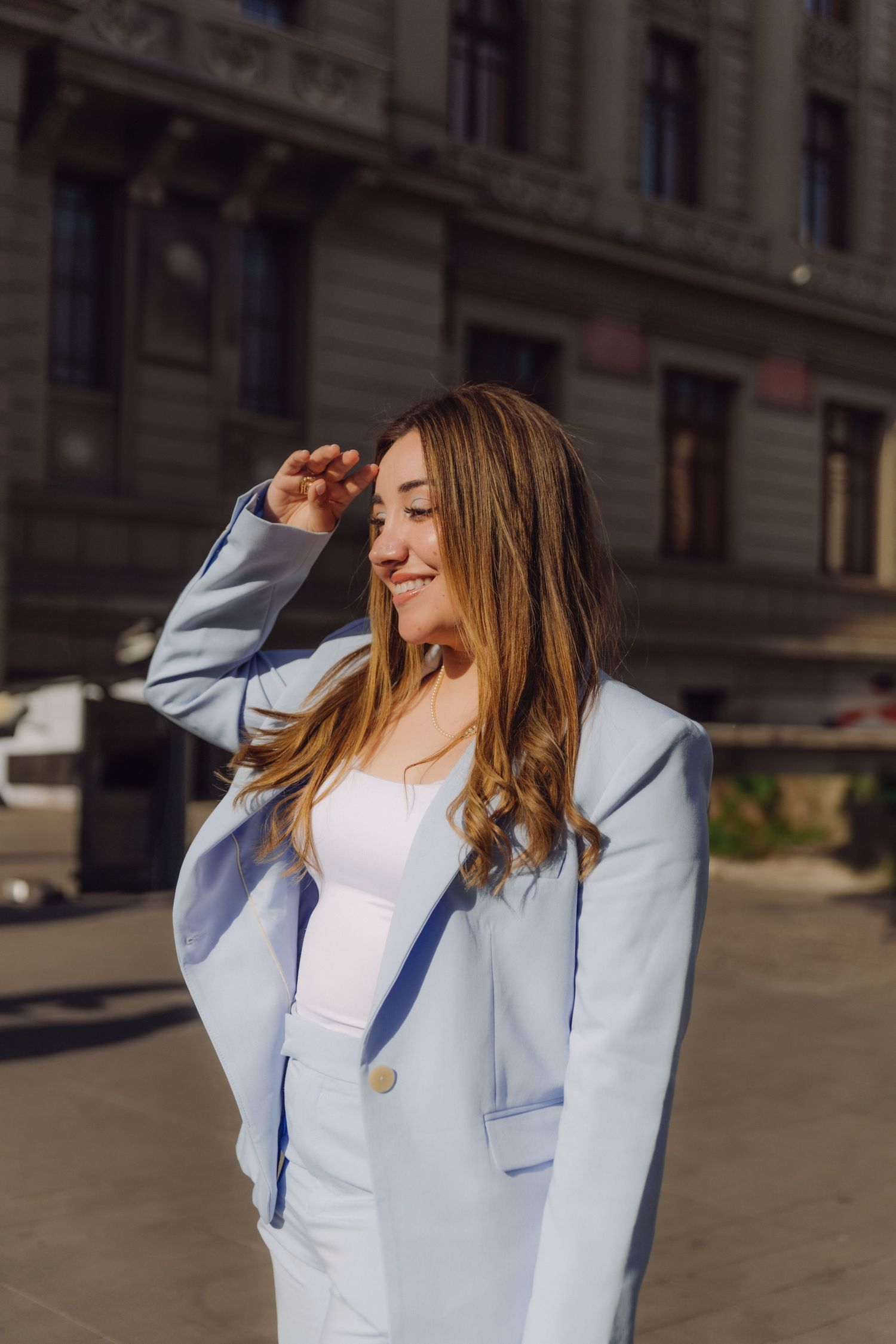 Smiling young woman in light blue suit shielding eyes from sun on urban street.