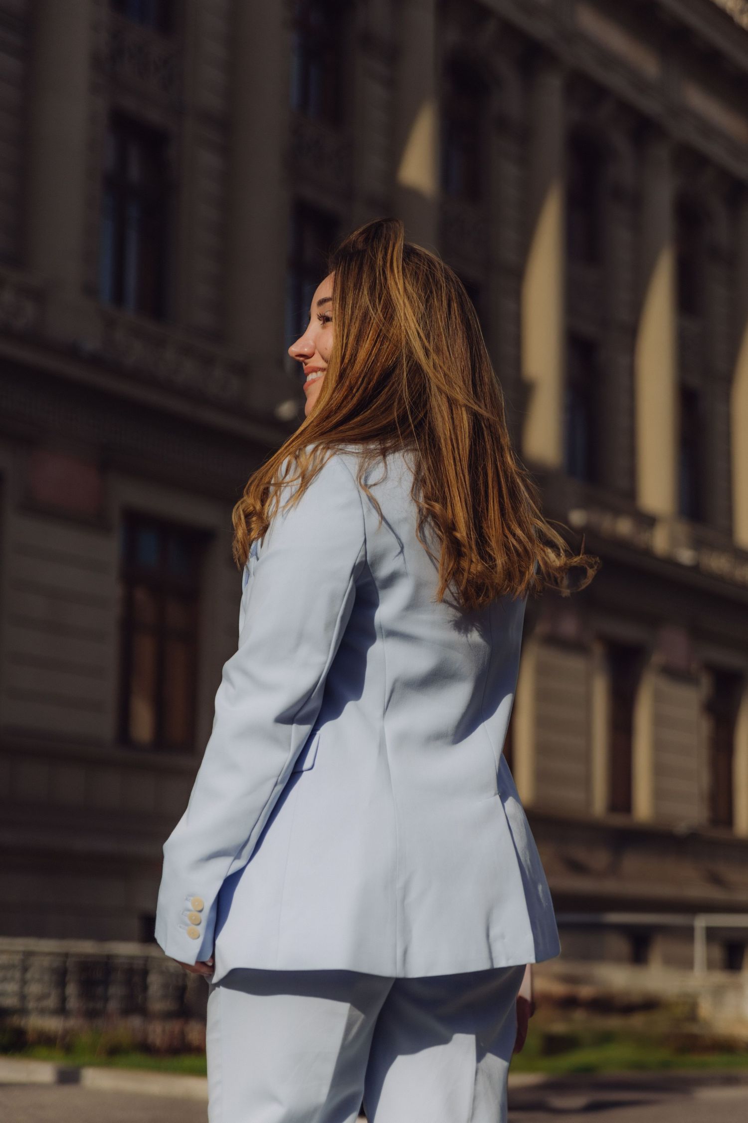 Young woman in light blue pantsuit viewed from behind on sunny city street.