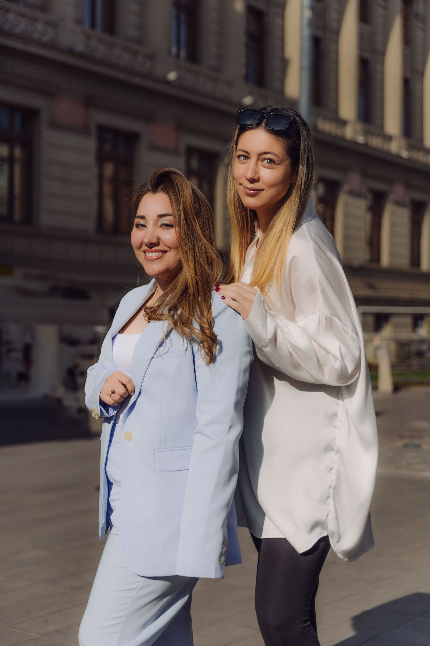 Two smiling women in blue and white blazers posing together on sunny city street.